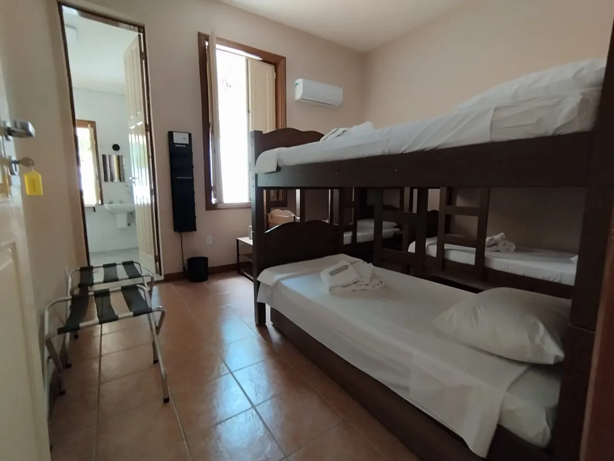 Quarto compartilhado com beliches de madeira, roupa de cama branca e banheiro privativo ao fundo.